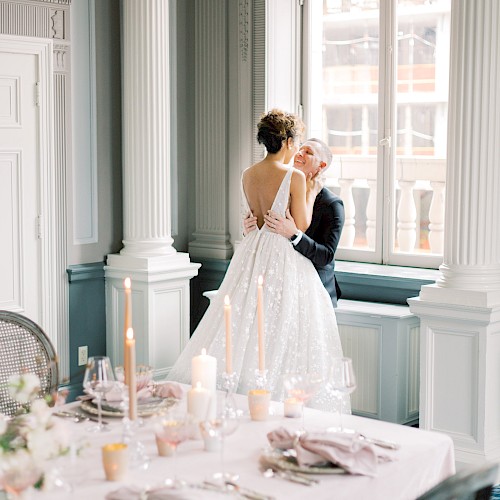 A bride and groom embrace near a window in a decorated room with a table set for a formal occasion, featuring candles and elegant tableware.