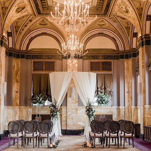 An elegant room set up for a wedding ceremony with chairs, floral arrangements, and draped fabric under ornate chandeliers and detailed ceilings.