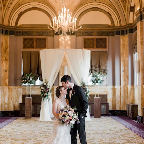 A couple is embracing in a lavish room, with ornate décor and chandeliers, in front of a floral wedding arch.