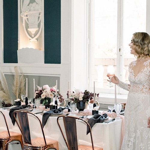 A woman in a white dress holds a glass, standing next to an elegantly set dining table with floral decorations and candles.