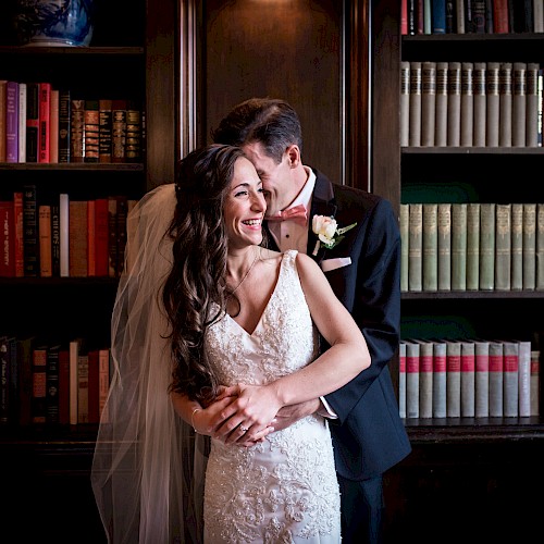 A couple in wedding attire is embracing in front of bookshelves, with the bride smiling and the groom leaning close, in a library setting.