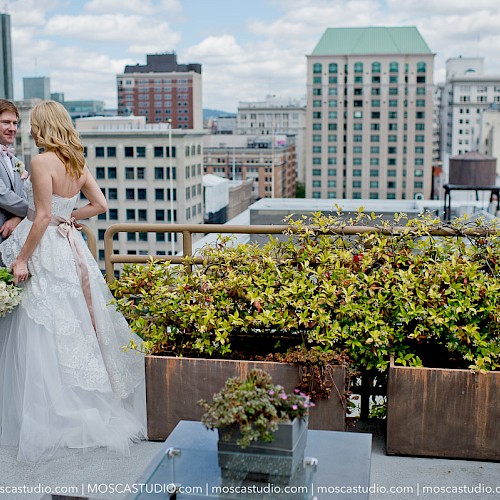 A couple is dressed in formal attire on a rooftop with city buildings in the background, surrounded by potted plants and foliage.