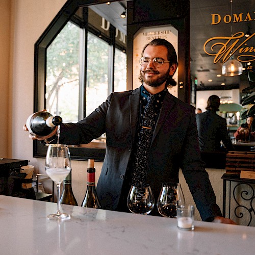 A man is pouring wine into a glass at a bar. There are three wine glasses and bottles on the counter. The setting is elegant and modern.