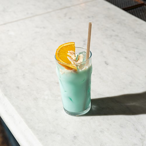 A light blue cocktail with ice, garnished with an orange slice and a paper straw, sits on a marble surface.