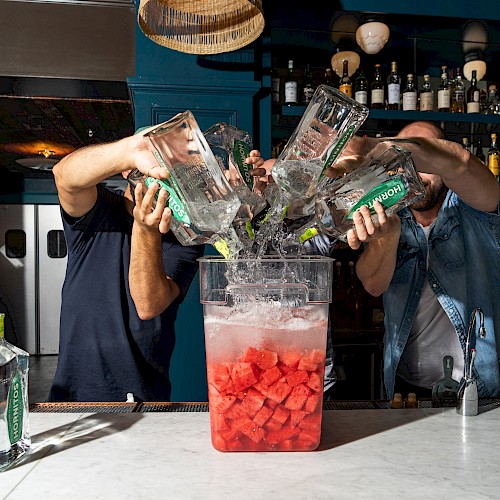 Three people are pouring bottles of gin into a container filled with ice and watermelon chunks on a bar counter.