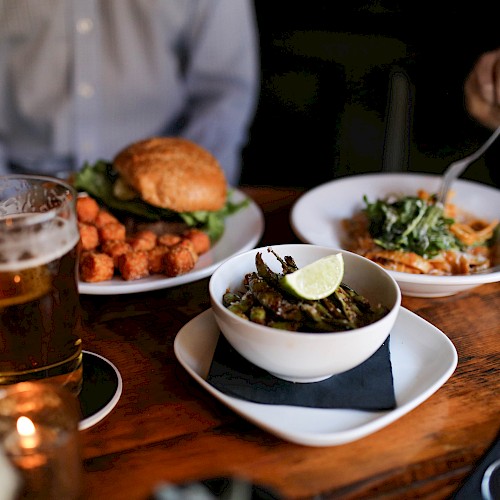 Two people dining with a burger, sweet potato fries, pasta, a bowl with lime, beer, and wine on a wooden table.