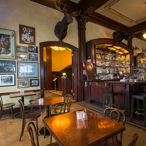 The image shows a cozy bar with wooden furniture, framed pictures on the wall, and mounted deer heads. The bar is stocked with various bottles.