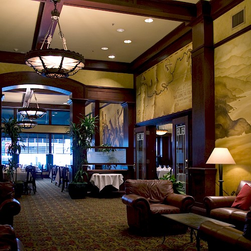 The image shows a dimly lit lounge with leather sofas, a patterned carpet, a large decorative mural, and an elegant chandelier.