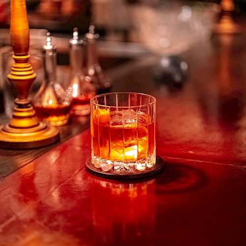 A glass of amber-colored beverage on a wooden bar counter, surrounded by dim lighting and decorative lamps.