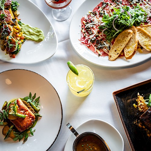 The image shows a table with gourmet dishes including steak, fish, carpaccio with bread, and colorful garnishes, alongside cocktails on a white tablecloth.