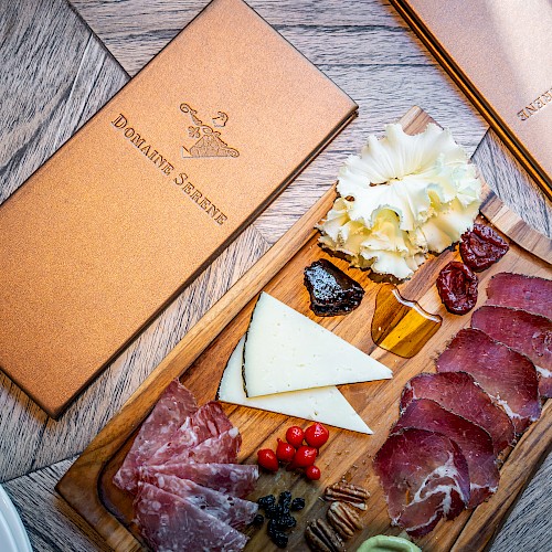 Charcuterie board with sliced meats, cheese, pickles, and garnish, next to leather-bound menus on a wooden table.