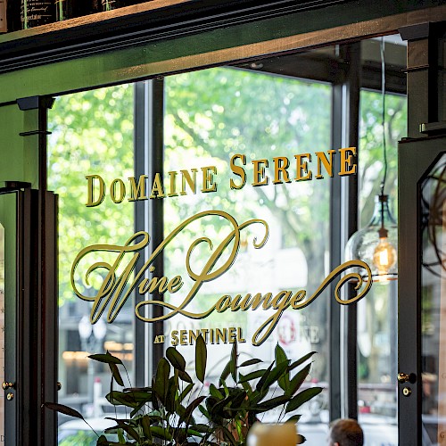 The image shows the Domaine Serene Wine Lounge with decorative plants, candles, and bottles, through a glass window with street view.