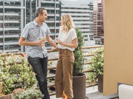 A couple is standing on a rooftop terrace, holding drinks and talking, with modern buildings in the background.