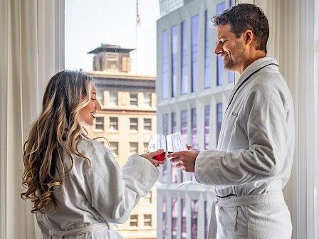 Two people in bathrobes clink glasses near a window with a city view, enjoying a relaxing moment.