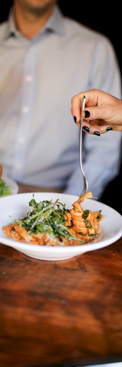 People sharing a meal; one has a burger, the other pasta with greens, and a drink is being held up for a toast.