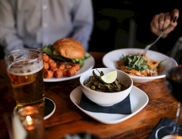 A table with a burger, salad, beer, red wine, and a bowl of lime-topped vegetables, with hands holding utensils.