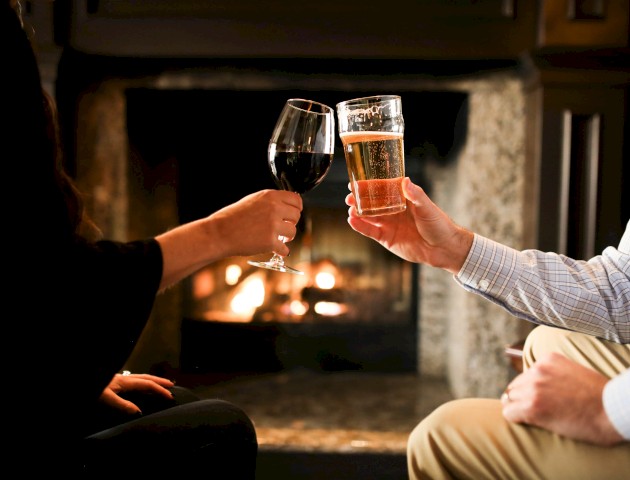 Two people are holding a wine glass and a beer glass while sitting by a fireplace, toasting each other in a cozy setting.