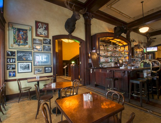 The image shows a cozy bar with wooden tables, chairs, a collection of wall photos, and mounted deer heads, creating a rustic atmosphere.