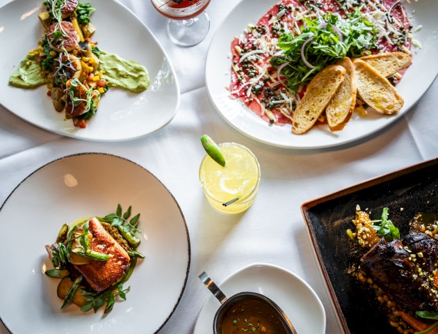 The image shows various gourmet dishes, including a steak, carpaccio, and salmon, served with drinks on a white tablecloth.