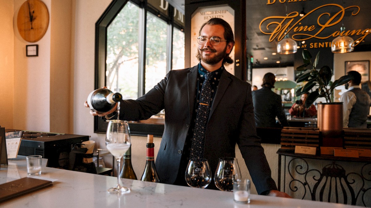 A person is pouring wine into a glass at a bar counter, with bottles and other glasses on the surface around them.