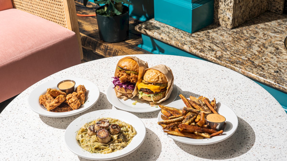 The image shows a table with two burgers, fries, pasta with vegetables, and fried appetizers, all served with dipping sauces.