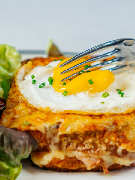 The image shows a person using a fork to cut into a croque madame with a fried egg, cheese, and ham, served with a side of leafy greens.