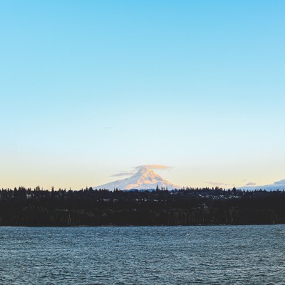 A distant snow-capped mountain under a blue sky, seen across a body of water with a treeline silhouette.