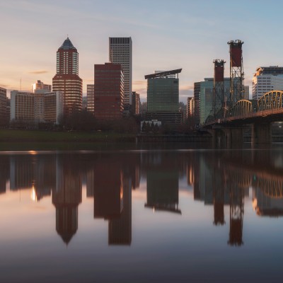 City skyline at sunset reflected in calm water, with a bridge on the right side of the image.