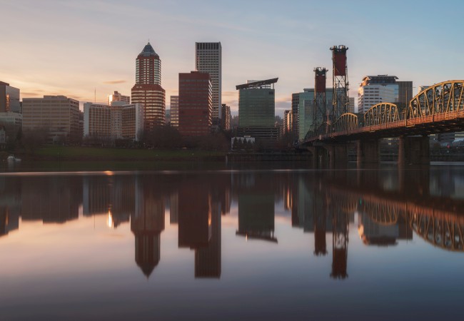 City skyline at sunset reflected in calm water, with a bridge on the right side of the image.