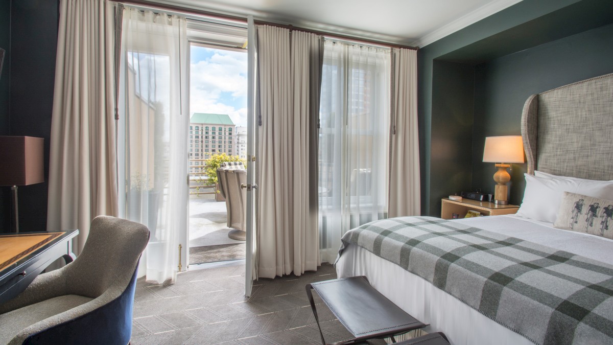 A cozy hotel room with a checkered bed, desk, and chair. It has a balcony with city views, allowing natural light through curtains.