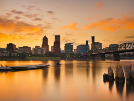 A city skyline at sunset with boats on calm river water, a bridge, and a few wooden posts, all bathed in warm orange and yellow tones.
