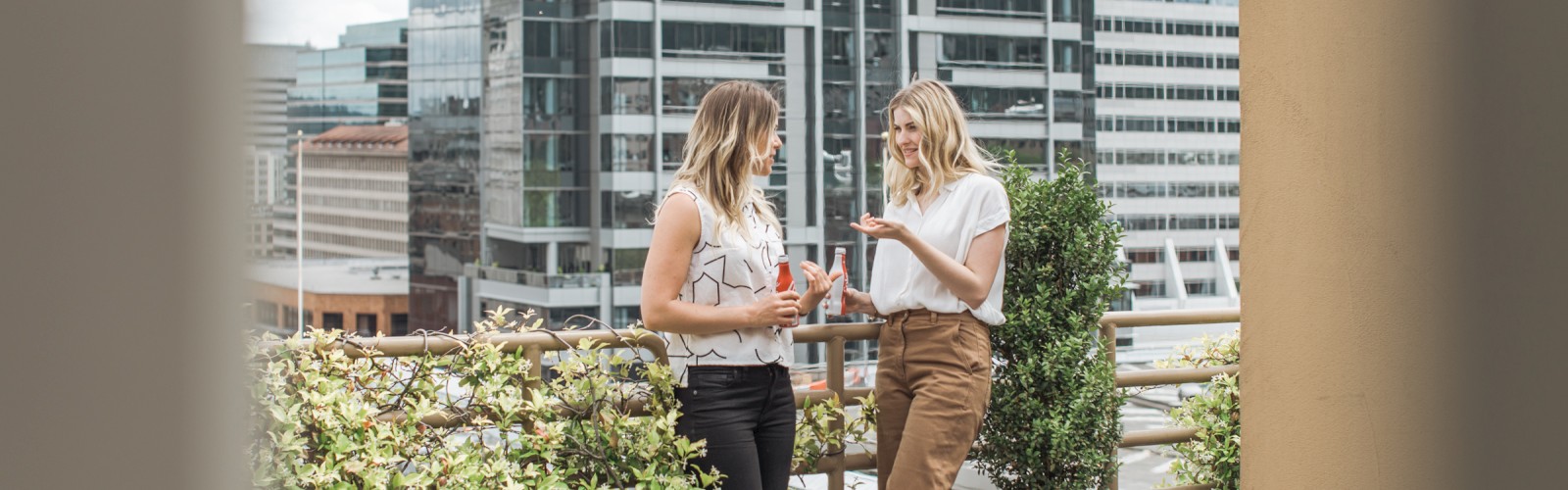 Two people chatting on a rooftop terrace in an urban setting, surrounded by plants and tall buildings in the background.