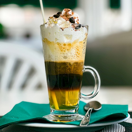 This image shows a layered coffee drink topped with whipped cream and chocolate chips, served in a glass on a saucer with a spoon.