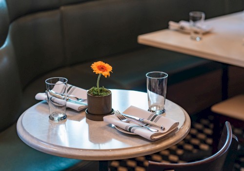 A small round table with place settings, glasses, and a potted flower in a cozy restaurant setting is shown.