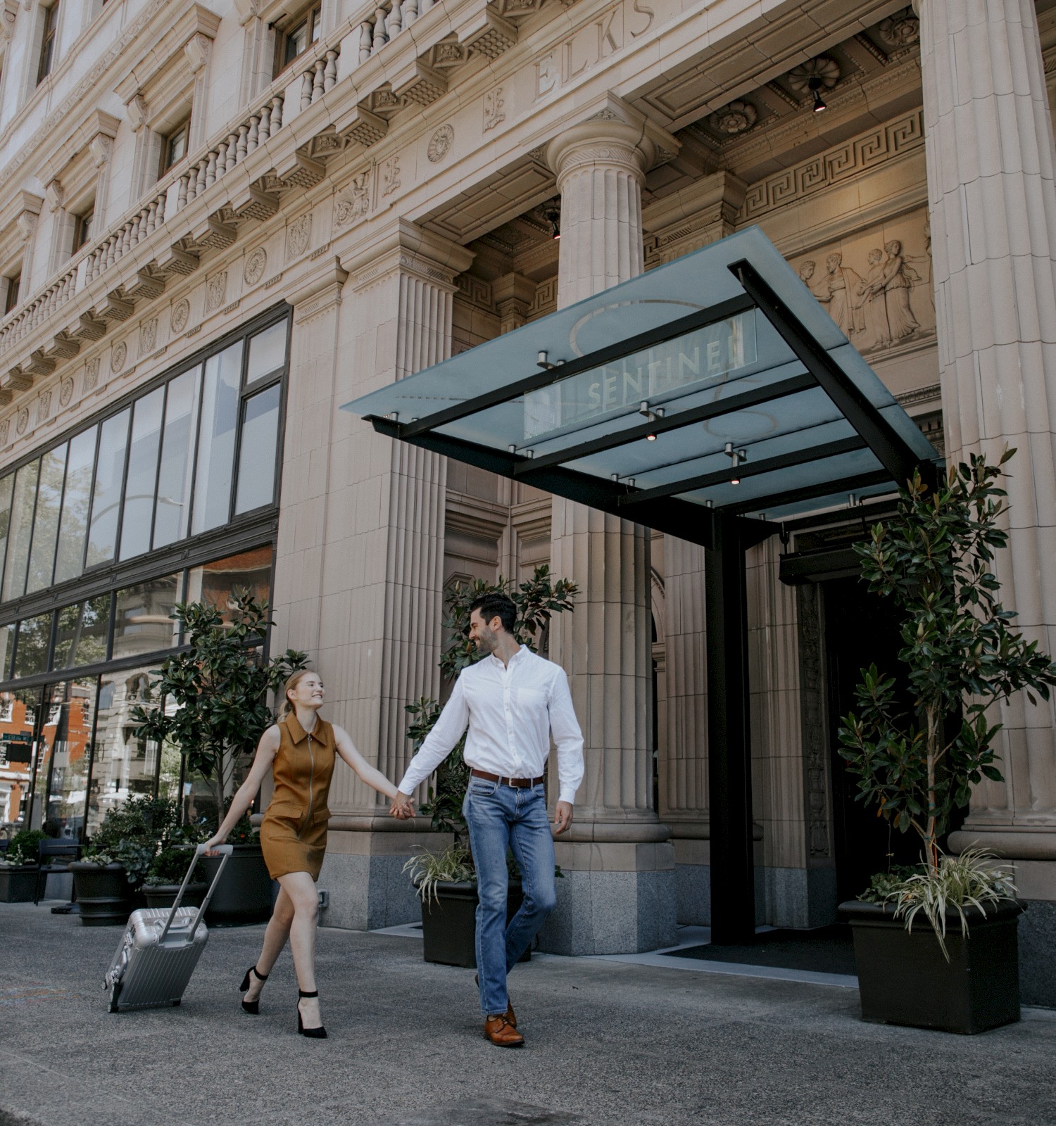 A couple exits a historic building, holding hands. One of them pulls a suitcase, suggesting travel or arrival.