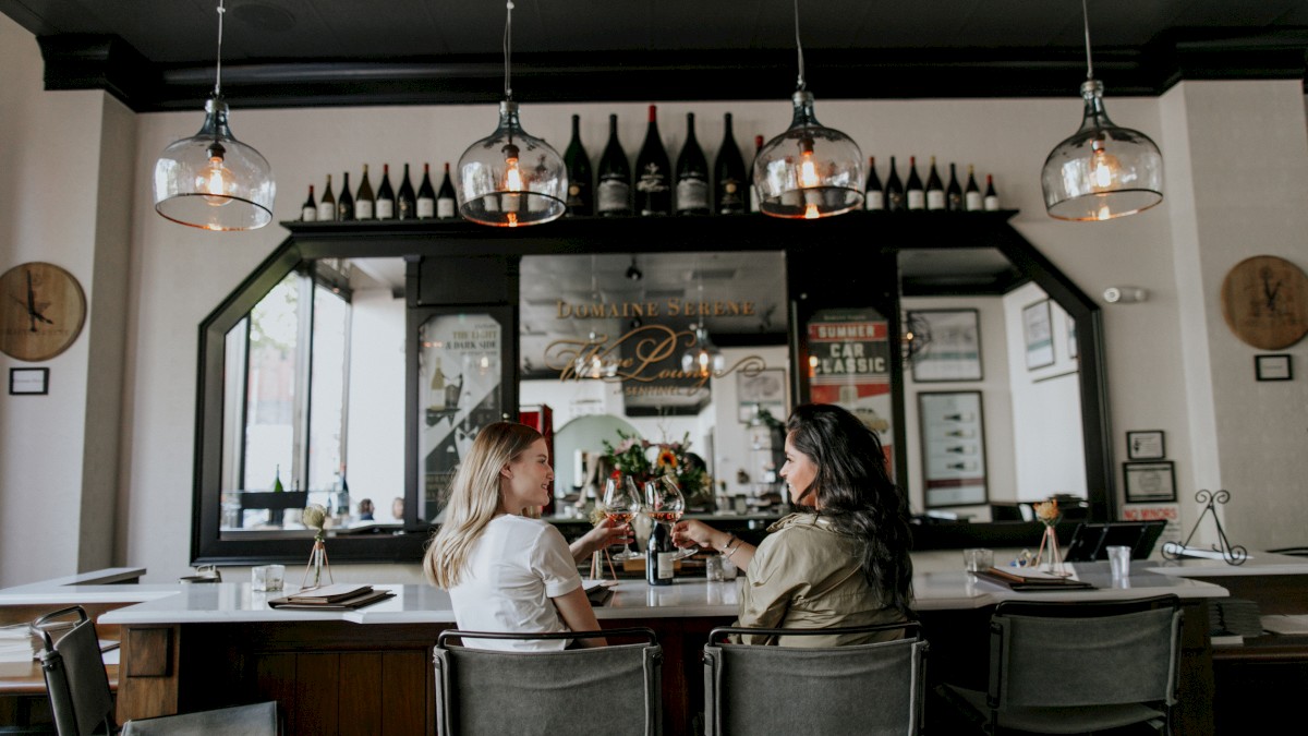 Two people are sitting at a bar, toasting with drinks. The bar is stylish with wine bottles and pendant lights overhead.