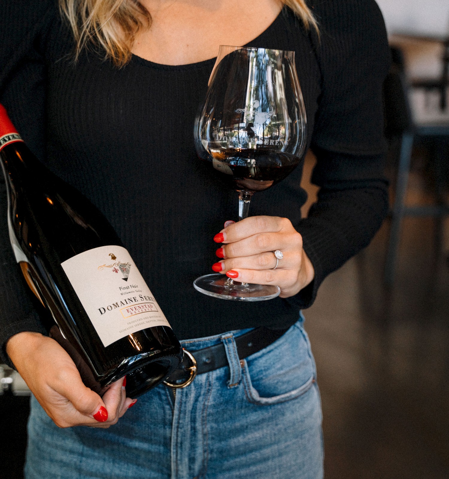 A person in a black top holds a bottle of red wine and a glass filled with wine, standing in what appears to be a restaurant.
