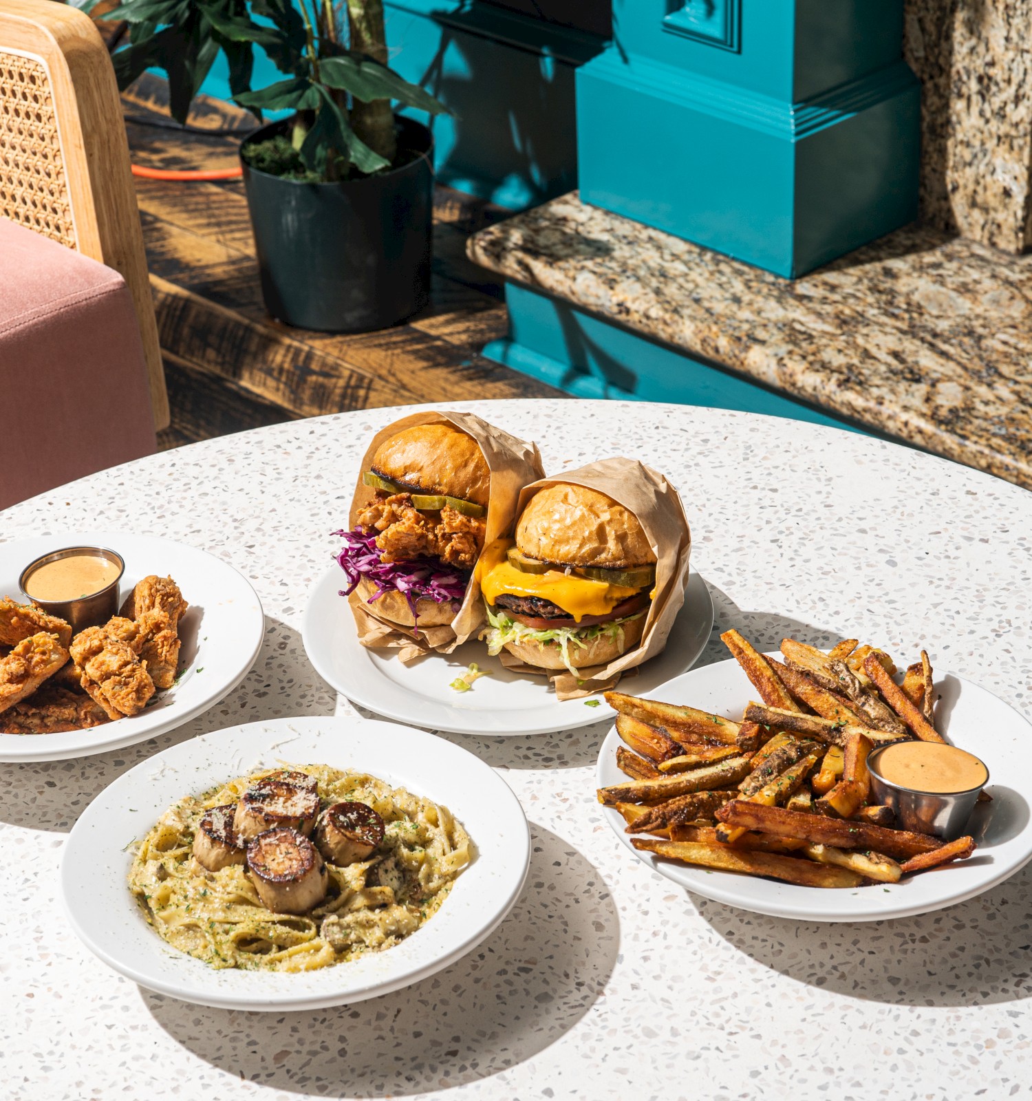 The image shows a table with fried chicken, burgers, fries, pasta, and dipping sauces in a cozy setting.