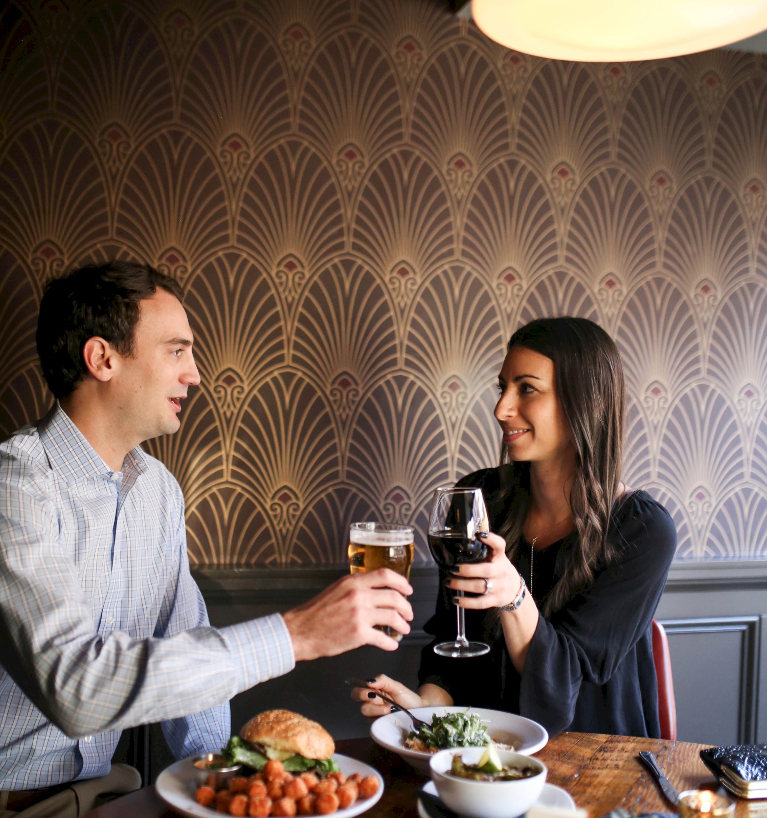 A couple toasting with drinks at a restaurant table with patterned wallpaper in the background.