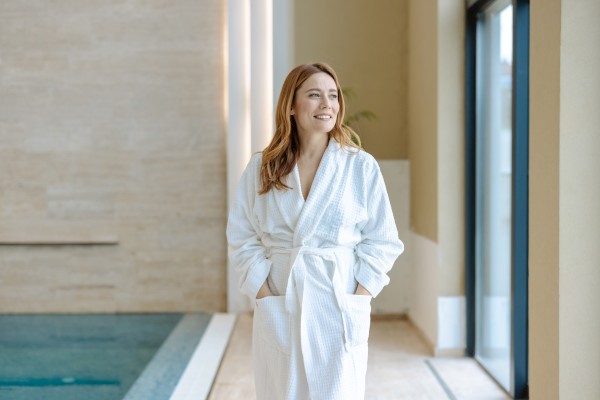 A person wearing a white robe stands indoors near a window, smiling, with their hands in the robe pockets, next to a pool.