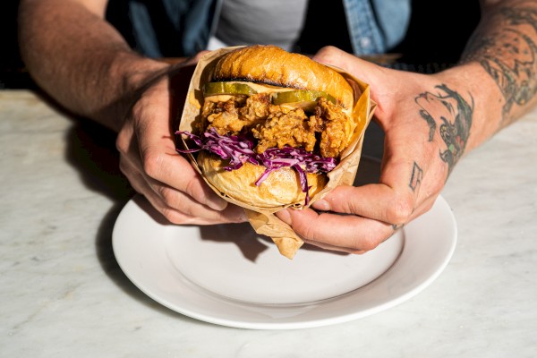 A person holds a sandwich with fried chicken, pickles, and purple coleslaw, wrapped in paper, over a white plate on a marble table.
