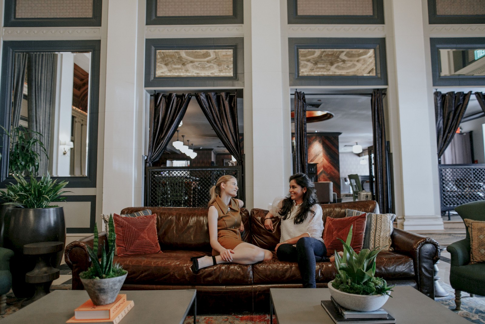 Two people are sitting on a brown leather couch, talking in a stylish room with plants and coffee tables in front of them.