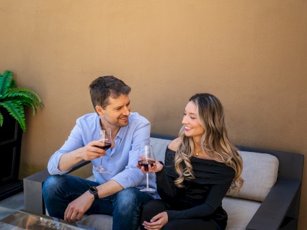 A couple is sitting on a patio sofa, toasting with wine glasses. Hanging lights and decorative plants are visible in the background.