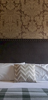 A neatly made bed with white pillows, a decorative pillow, and a checkered blanket in a warmly lit bedroom with floral wallpaper.