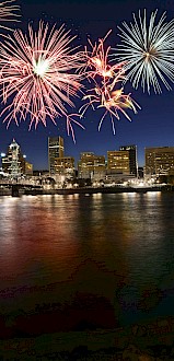 Fireworks light up the night sky over a city skyline reflected in a river.