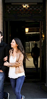 A smiling man and woman walk arm-in-arm out of a building entrance, enjoying each other's company.