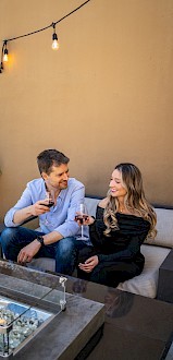 A couple sitting on a patio couch, clinking wine glasses, with hanging lights and a fire pit in the foreground.