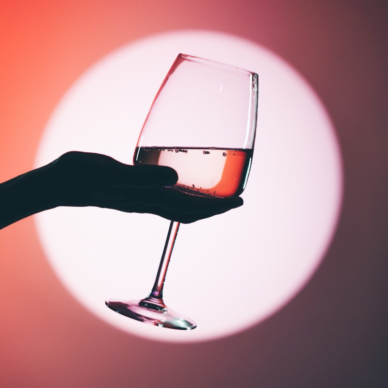 A hand holds a wine glass up against a warm, pinkish-red circular light, as if to toast or admire a drink.