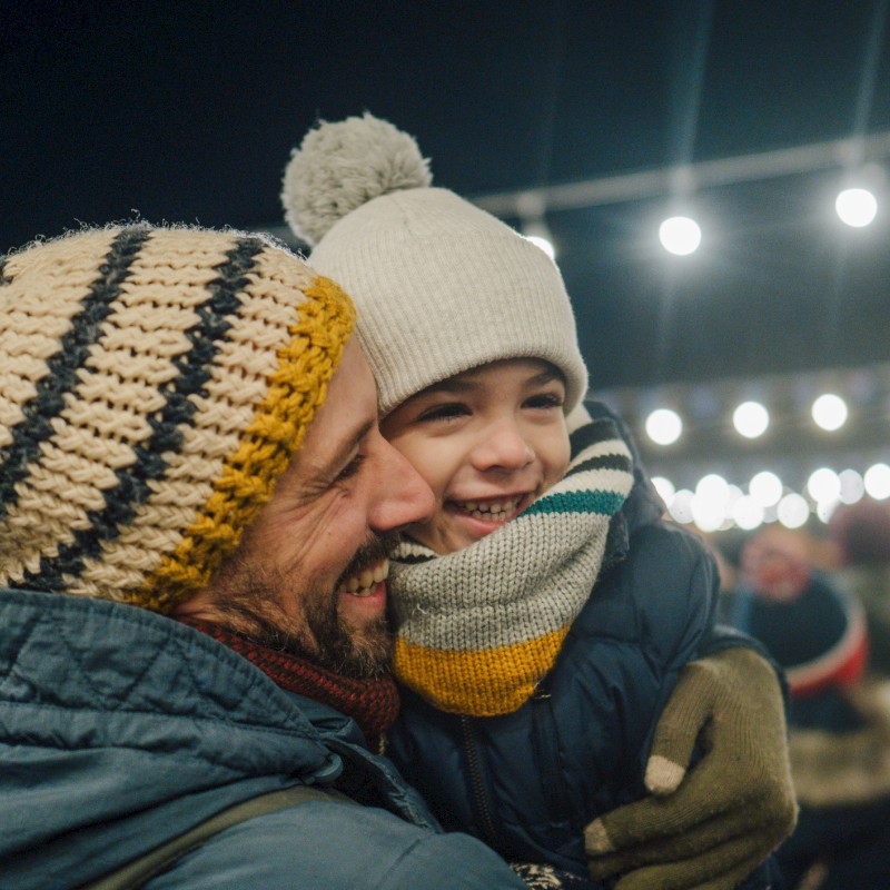 Two joyful people sharing a hug at a festive night market, bundled in warm knit hats, scarves, and cozy jackets under string lights.