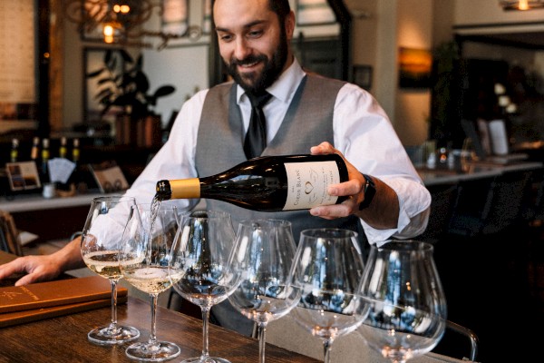 A man is pouring wine into multiple glasses on a bar counter, with bottles and lights in the background, creating a cozy atmosphere.